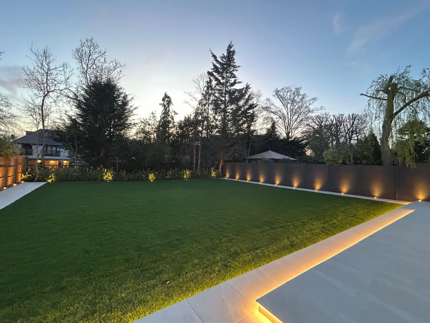 Finished rear garden with paved paths, level lawn and warm perimeter lighting at sunset